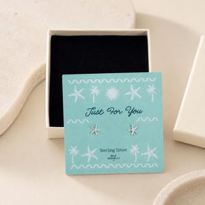 Pode incluir: Um par de brincos de tarracha em forma de estrela-do-mar em prata de lei, apresentados num cartão azul-claro com ilustrações brancas de palmeiras e sol. O cartão apresenta o texto Just For You e Sterling Silver dentro de uma caixa branca.
