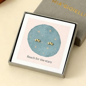Può includere: Una scatola di gioielli grigia con il testo dorato "MIA GIOIELLI" sul coperchio. All'interno, una carta mostra orecchini a forma di stella dorata. La carta ha un cerchio blu con stelle dorate e il testo "Reach for the stars".