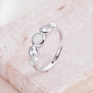 May include: A silver ring with a row of alternating clear and opalescent stones. The adjustable ring has a delicate band. The design is simple and elegant, suitable for everyday wear. The ring is set against a soft, neutral background. The stones are round.