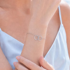 Op de afbeelding: Een delicate zilverkleurige kettingarmband met twee in elkaar grijpende cirkelvormige ringen als centraal middelpunt. De ene ring heeft een gladde, gepolijste afwerking, terwijl de andere ring is versierd met kleine, heldere, fonkelende steentjes.