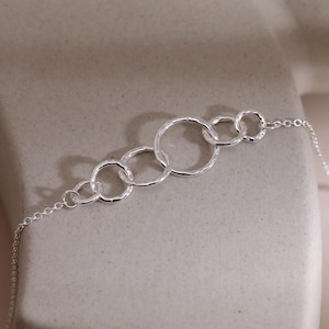 May include: A silver bracelet featuring a series of interconnected hammered rings. The rings vary in size and are linked together in a linear design. The bracelet has a delicate chain and clasp.