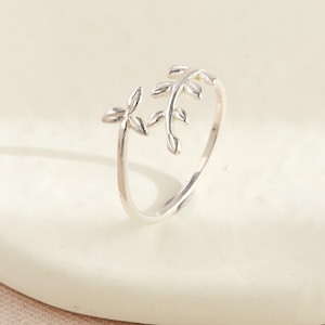 May include: A silver ring with a leaf design. The ring has two leaf clusters, one on each side of the band, creating an open design. The leaves have detailed veining, adding to the ring's natural aesthetic. The ring is displayed on a white surface.