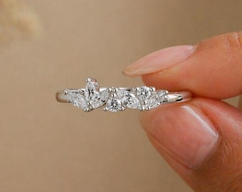 May include: A delicate silver ring featuring a cluster of clear, sparkling gemstones. The ring's design includes a mix of shapes, with a pear-cut stone at the center and other stones on either side. The ring is held up by a hand.