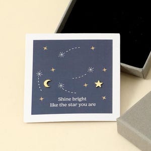 Könnte beinhalten: Eine graue Schmuckschatulle mit dem goldenen Text "MIA GIOIELLI" ist geöffnet und zeigt einen schwarzen Innenraum. Eine kleine Karte mit einem Nachthimmel-Design und dem Text "Shine bright like the star you are" liegt neben der Schachtel. Die Schachtel steht auf einer hellen Oberfläche.