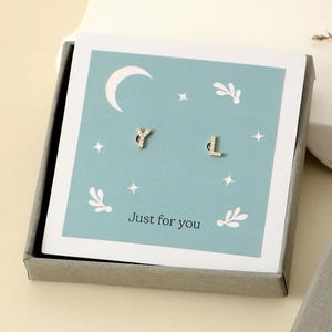 Könnte beinhalten: Goldfarbene Buchstabenohrringe in Form von "Y" und "D", präsentiert in einer Geschenkbox. Die Box enthält eine hellblaue Karte mit Mond, Sternen und dem Schriftzug "Just for you". Die äußere Box ist grau mit goldfarbenen Buchstaben.