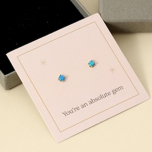 Könnte beinhalten: Ein Paar kleiner, runder Ohrstecker mit blauen Edelsteinen auf einer rosa Karte mit dem Text "You're an absolute gem". Die Ohrringe werden in einer offenen grauen Schachtel mit schwarzem Innenraum neben einer geschlossenen grauen Schachtel mit goldenem Text "MIA GIOIELLI" präsentiert.