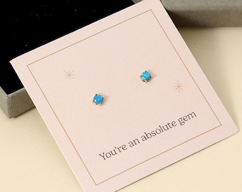 Può includere: Un paio di piccoli orecchini a bottone rotondi con pietre preziose blu su una carta rosa con la scritta "You're an absolute gem". Gli orecchini sono esposti in una scatola grigia aperta con interno nero, accanto a una scatola grigia chiusa con testo dorato "MIA GIOIELLI."