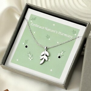 May include: A silver leaf pendant necklace displayed in an open gray gift box with the text "Embrace Nature's Harmony" on a light green background. The box is next to a closed gray box with gold text that reads "MIA GIOIELLI".