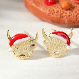 Könnte beinhalten: Ein Paar goldfarbene Ohrstecker mit einem Highland-Kuh-Design. Jede Kuh trägt einen rot-weißen Weihnachtsmannhut. Die Ohrringe liegen auf einem weißen Teller, im Hintergrund ein Lebkuchen.
