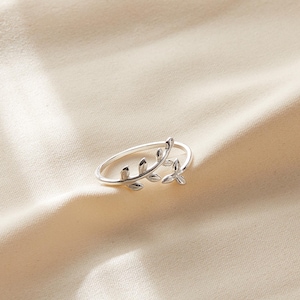 May include: A silver-colored ring with a delicate leaf design. The ring features a thin band and a detailed leaf motif that wraps around the finger. The leaves have a textured appearance, adding depth to the jewelry.