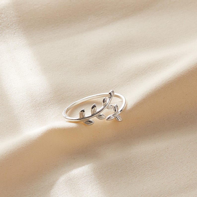 May include: A silver-colored ring with a delicate leaf design. The ring features a thin band and a detailed leaf motif that wraps around the finger. The leaves have a textured appearance, adding depth to the jewelry.
