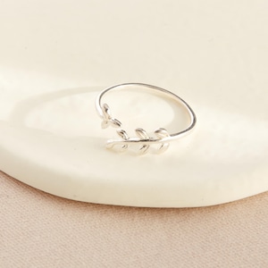 May include: A silver-colored ring with a leaf design. The ring features a delicate band that wraps around the finger, with a detailed leaf motif on the front. The ring is shown on a white surface.