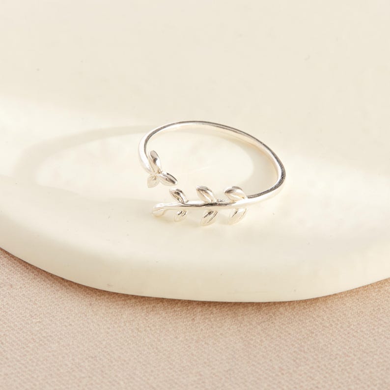 May include: A silver-colored ring with a leaf design. The ring features a delicate band that wraps around the finger, with a detailed leaf motif on the front. The ring is shown on a white surface.