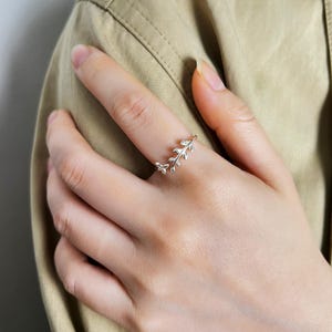 May include: A silver-colored ring with a delicate leaf design. The ring features a small, open design with a branch of leaves wrapping around the finger. The ring is worn on a person's finger, against a neutral background.