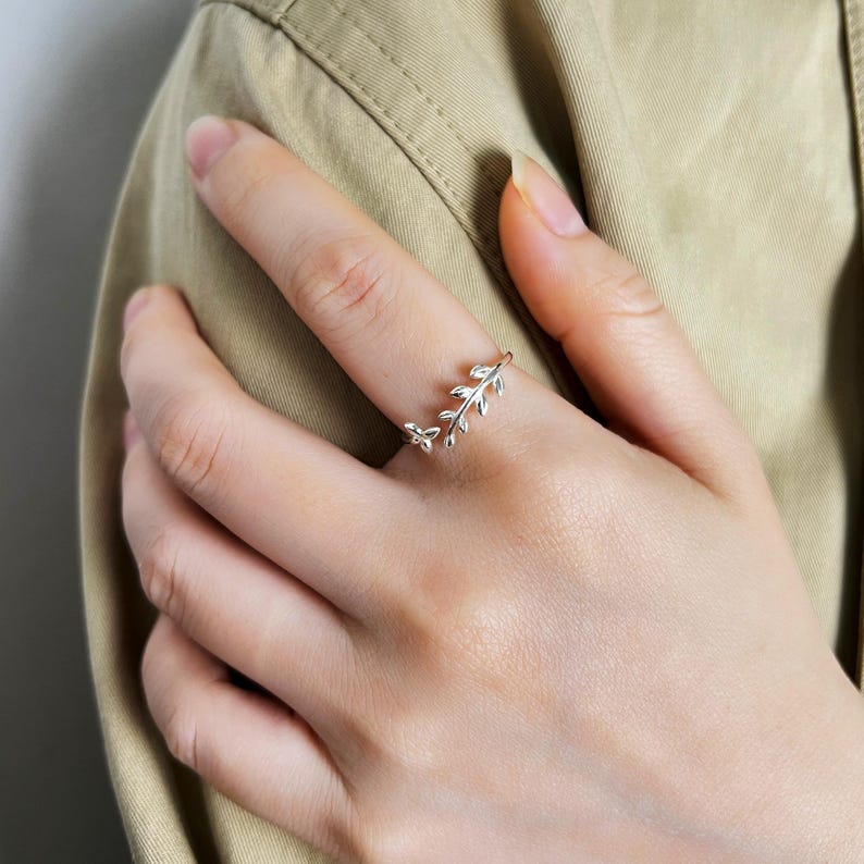 May include: A silver-colored ring with a delicate leaf design. The ring features a small, open design with a branch of leaves wrapping around the finger. The ring is worn on a person's finger, against a neutral background.
