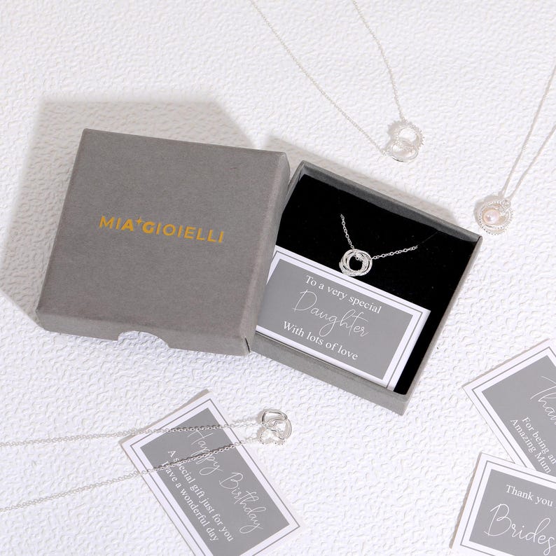Pu&ograve; includere: Una collana in argento con ciondolo circolare all'interno di una scatola regalo grigia con l'etichetta MIA GIOIELLI. La scatola include un biglietto con la scritta To a very special Daughter With lots of love. Altre collane e biglietti sono visibili.