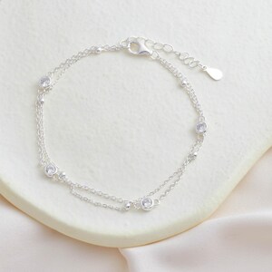 Puede incluir: Un delicado brazalete de plata con un dise&ntilde;o de cadena doble y peque&ntilde;as piedras de circonita c&uacute;bica transparentes.