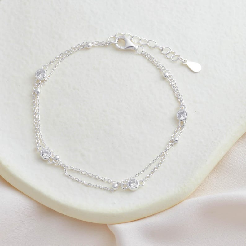 Puede incluir: Un delicado brazalete de plata con un dise&ntilde;o de cadena doble y peque&ntilde;as piedras de circonita c&uacute;bica transparentes.