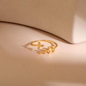 May include: A gold-colored ring with a leaf design. The ring features a delicate band that wraps around the finger, with a small leaf and a larger leaf cluster on the top. The ring is set against a neutral background.