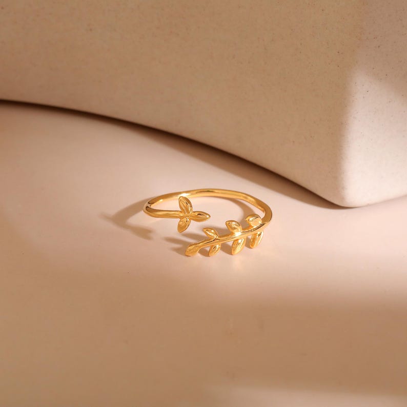May include: A gold-colored ring with a leaf design. The ring features a delicate band that wraps around the finger, with a small leaf and a larger leaf cluster on the top. The ring is set against a neutral background.
