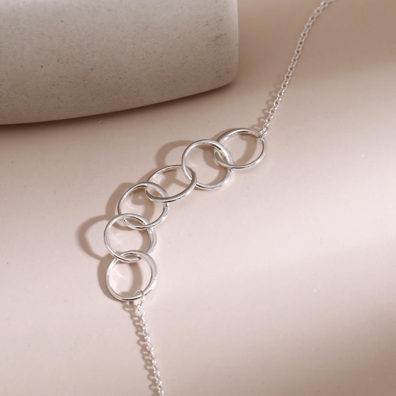 Pu&ograve; includere: Una collana in argento con un punto focale centrale composto da cinque anelli circolari interconnessi. La catena &egrave; sottile e delicata, con una finitura metallica lucida che riflette la luce, creando un aspetto semplice e pulito.