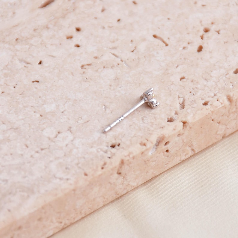May include: A single silver stud earring with a small, clear gemstone cluster. The earring is set against a light beige, textured surface. The earring post is visible, showing the details of the jewelry.