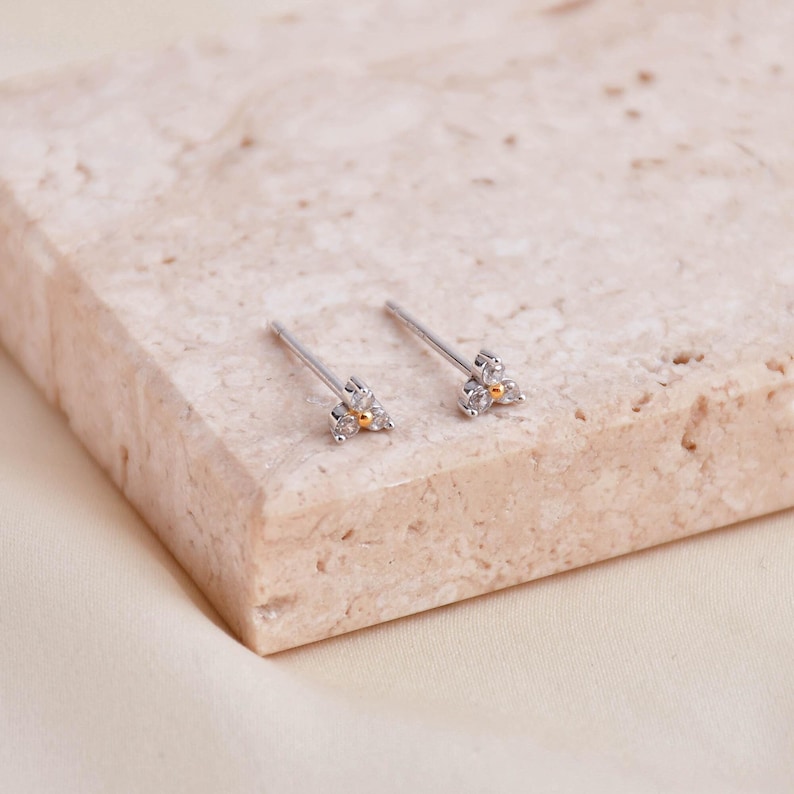 May include: A pair of delicate silver stud earrings. Each earring features a small, three-petal design with clear gemstones and a gold-colored center. The earrings are displayed on a light-colored, textured surface.