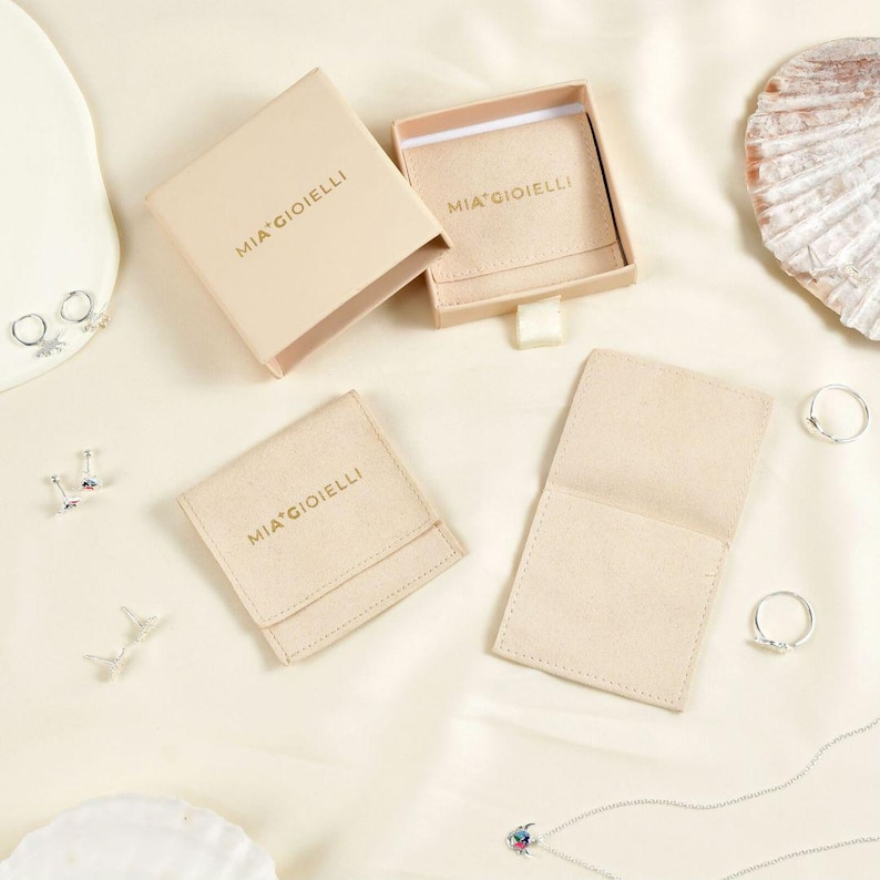 May include: An overhead shot of jewelry packaging and accessories. The image features open and closed beige boxes and pouches with the text "MIA GIOIELLI" printed in gold. Several pieces of jewelry, including rings and earrings, are scattered around the packaging.