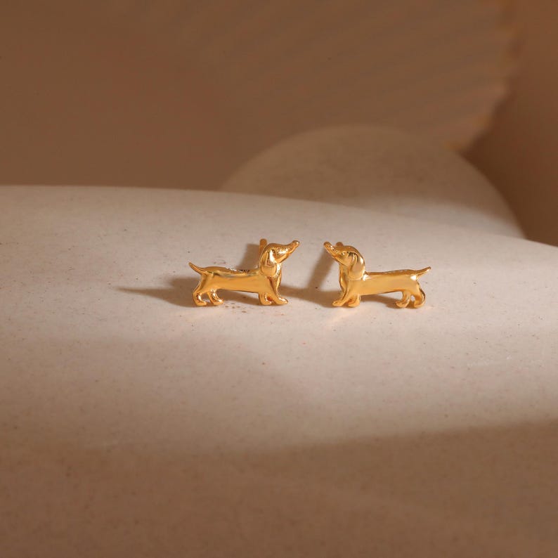 Op de afbeelding: Twee goudkleurige teckel oorknopjes. De oorbellen zijn gevormd als kleine honden, met gedetailleerde kenmerken. Ze worden gepresenteerd op een neutraal oppervlak, met zachte verlichting die schaduwen cre&euml;ert.