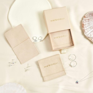 May include: An overhead shot of jewelry and packaging. The image features a light pink box and pouches with the text "MIA GIOIELLI" in gold. There are also silver earrings, rings, and a necklace with a colorful pendant.