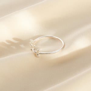 May include: A silver ring with a delicate floral design. The ring features a thin band and small, stylized flowers clustered on one side. The ring is displayed on a cream-colored fabric.