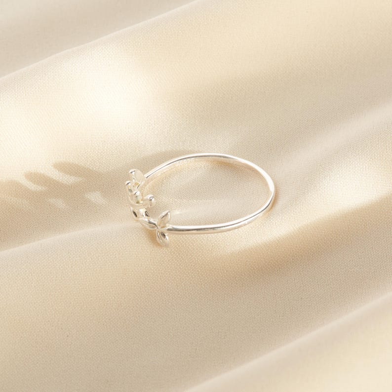 May include: A silver ring with a delicate floral design. The ring features a thin band and small, stylized flowers clustered on one side. The ring is displayed on a cream-colored fabric.