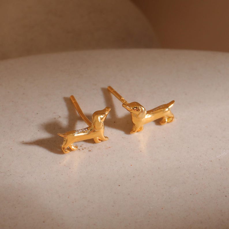 Op de afbeelding: Een paar goudkleurige teckel oorknopjes. Elke oorbel is een miniatuursculptuur van een teckel, met gedetailleerde kenmerken. De oorbellen staan tegen een neutrale achtergrond, wat hun metallic glans benadrukt.