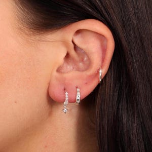 Puede incluir: Primer plano de la oreja de una persona con tres pendientes de plata. El pendiente inferior es un pendiente de estrella colgante con pequeñas piedras transparentes. El pendiente del medio es un pequeño aro de plata. El pendiente superior es un aro de plata más grande.