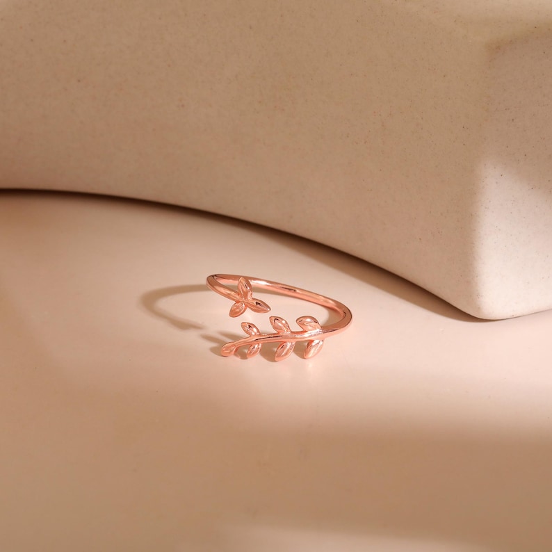 May include: A rose gold ring with a leaf design. The ring features a delicate open band with a small leaf at the top and a branch of leaves curving around the finger. The ring is displayed on a light pink surface.