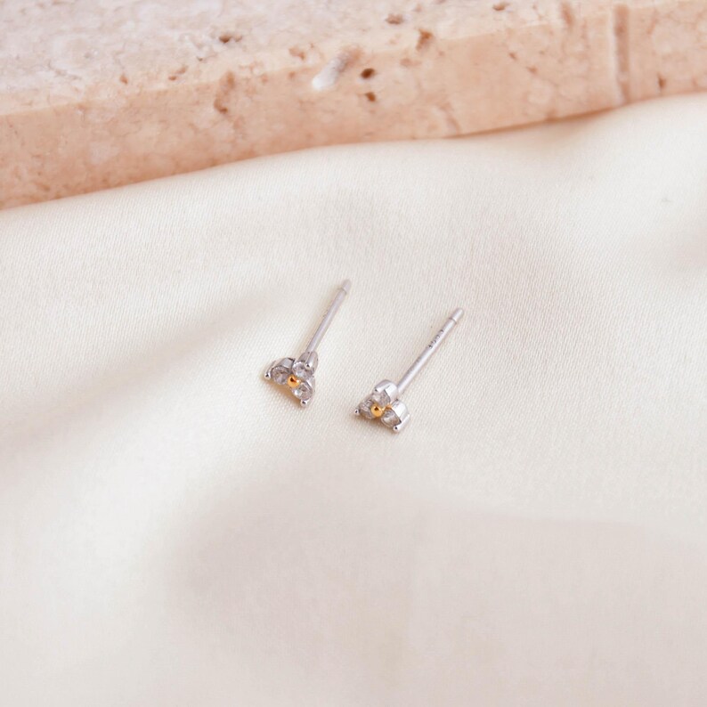 May include: Two silver stud earrings with a triangular design. Each earring features three clear stones and a small gold-colored accent. The earrings are displayed on a white, textured surface.