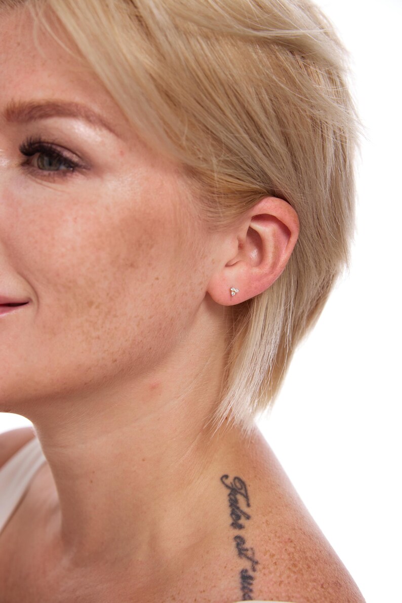 May include: A close-up of a person's ear with a small, delicate stud earring. The earring has a subtle, sparkling design. The person has short, blonde hair and a tattoo on their shoulder.