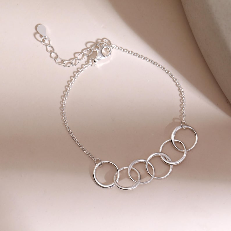 Pu&ograve; includere: Un braccialetto d'argento con sei cerchi interconnessi. Il braccialetto ha una catena delicata e una chiusura regolabile. I cerchi sono uniformemente distanziati, creando un design moderno e minimalista. Il braccialetto &egrave; esposto su una superficie rosa chiaro.