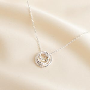 May include: A silver-coloured necklace featuring a pendant composed of three interconnected, irregular circular rings of varying sizes. The pendant hangs from a delicate, thin silver-coloured chain, creating a layered and textured appearance.