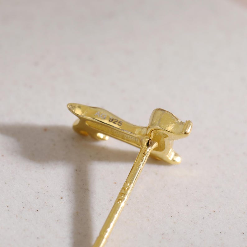 Op de afbeelding: Close-up van een goudkleurige oorbel met een pin. De oorbel heeft een gestileerde vorm met een getextureerd oppervlak. De markering "925" is zichtbaar op de achterkant van de oorbel.