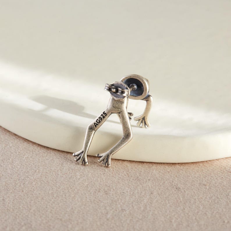May include: Silver frog earring with a post back. The back of the earring is stamped with the text "AG925".