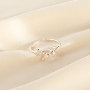 May include: A silver-colored ring with a delicate leaf design. The ring features a slender band and a detailed leaf motif, adding a touch of nature-inspired elegance. The ring is set against a soft, cream-colored background.
