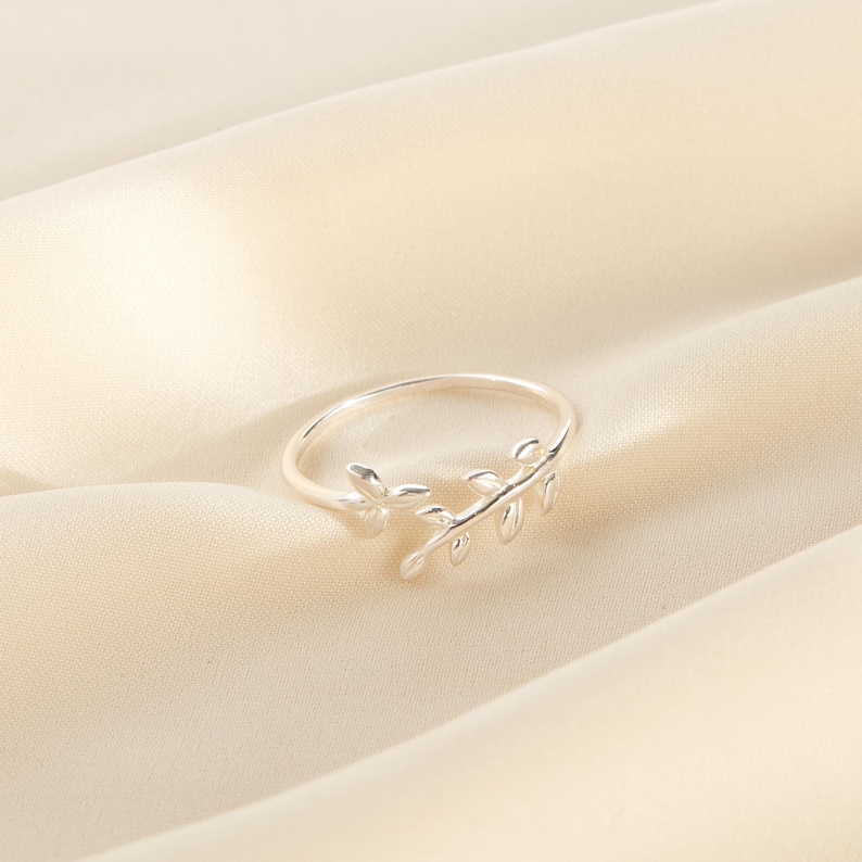 May include: A silver-colored ring with a delicate leaf design. The ring features a slender band and a detailed leaf motif, adding a touch of nature-inspired elegance. The ring is set against a soft, cream-colored background.