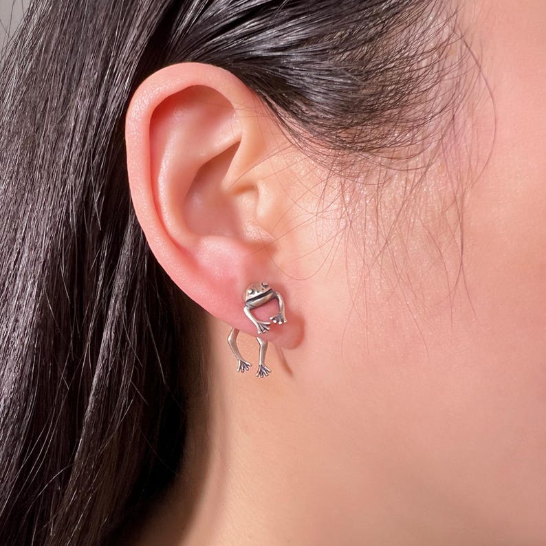 May include: Silver frog earrings with a dangling design. The frog is sitting on a small platform with its legs dangling down.