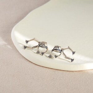 May include: A pair of silver frog earrings. The frogs are facing forward with their legs outstretched.