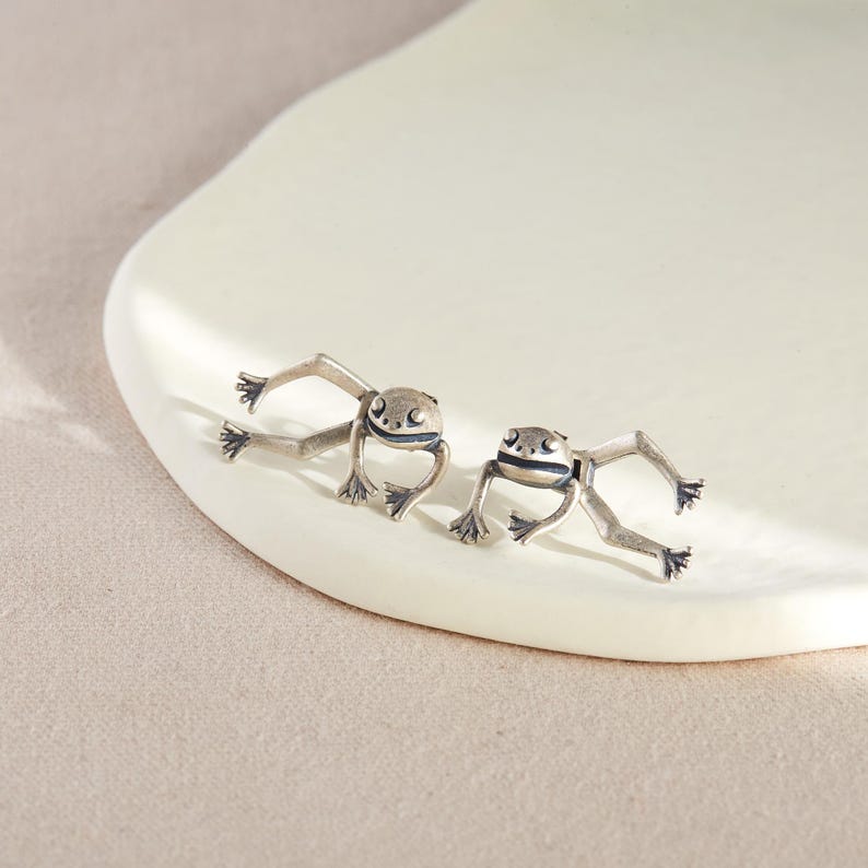 May include: A pair of silver frog earrings. The frogs are facing forward with their legs outstretched.