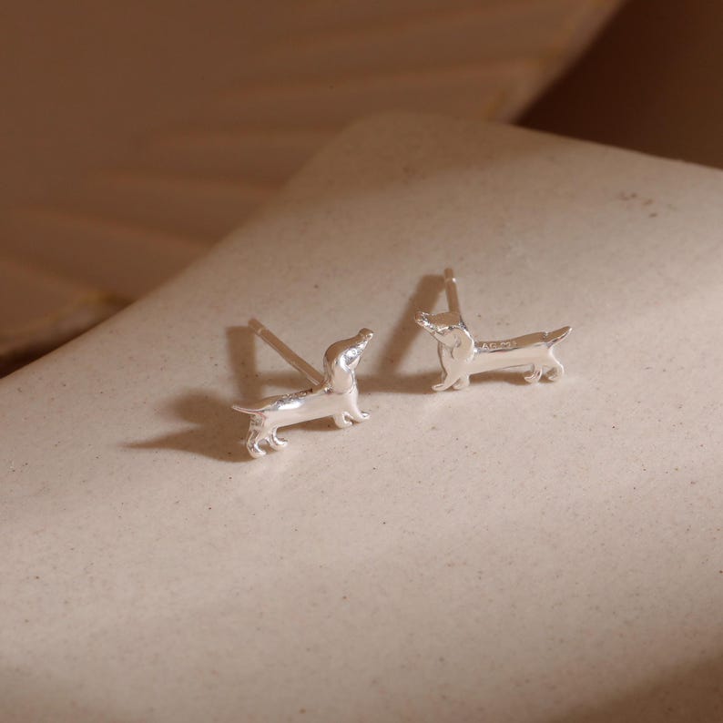 Op de afbeelding: Een paar zilveren teckel oorknopjes. Elke oorbel toont een gedetailleerde miniatuur teckel met een pin voor doorboorde oren. De oorbellen worden gepresenteerd op een neutrale ondergrond, waarbij hun gepolijste afwerking en ontwerp worden benadrukt.