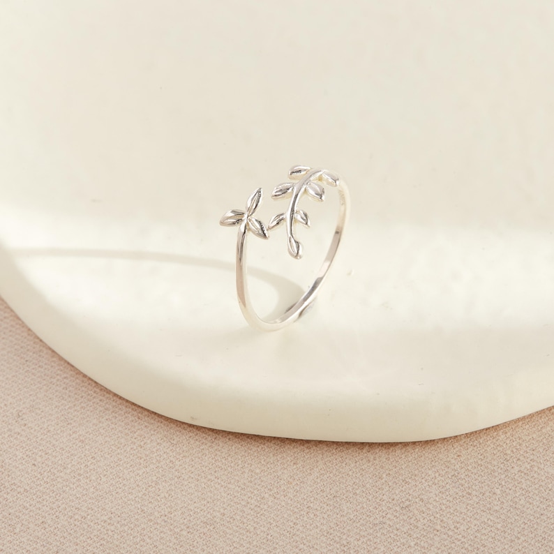 May include: A silver-colored ring with a delicate leaf design. The ring features two leaf clusters, one on each side of the band, creating an open design. The leaves have detailed veining, adding to the ring's natural aesthetic. The ring is displayed on a white surface.
