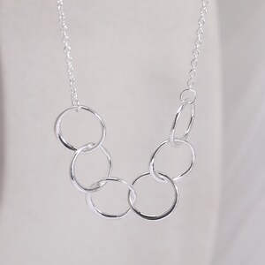 Pu&ograve; includere: Una collana color argento con una catenina sottile e un ciondolo composto da cinque anelli circolari intrecciati di dimensioni diverse, disposti in una forma curva e a cascata che pende simmetricamente dai collegamenti della catena.