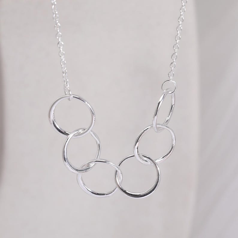 Pu&ograve; includere: Una collana color argento con una catenina sottile e un ciondolo composto da cinque anelli circolari intrecciati di dimensioni diverse, disposti in una forma curva e a cascata che pende simmetricamente dai collegamenti della catena.